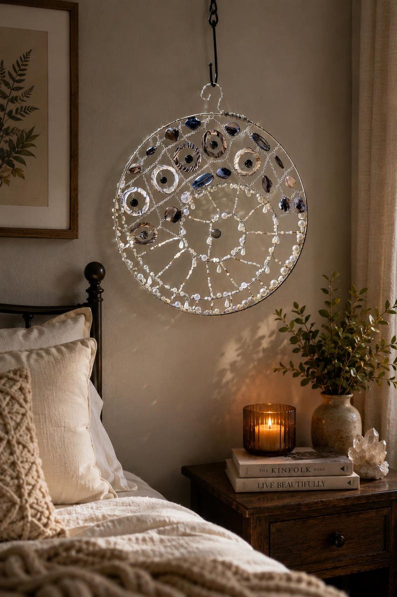 May include: A circular wall hanging featuring a silver wire frame adorned with dark blue, grey, and clear glass beads arranged in a spiral pattern. The piece is suspended from a black metal hook and incorporates various bead shapes and sizes.