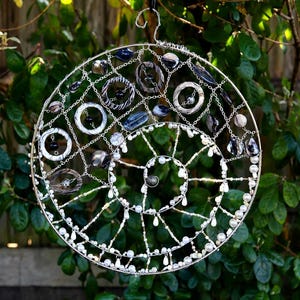 May include: A circular wire wall hanging featuring a spiral design. The structure is adorned with various white, grey, and black beads, along with several circular rings and dark stone-like accents integrated into the silver-toned wire frame.