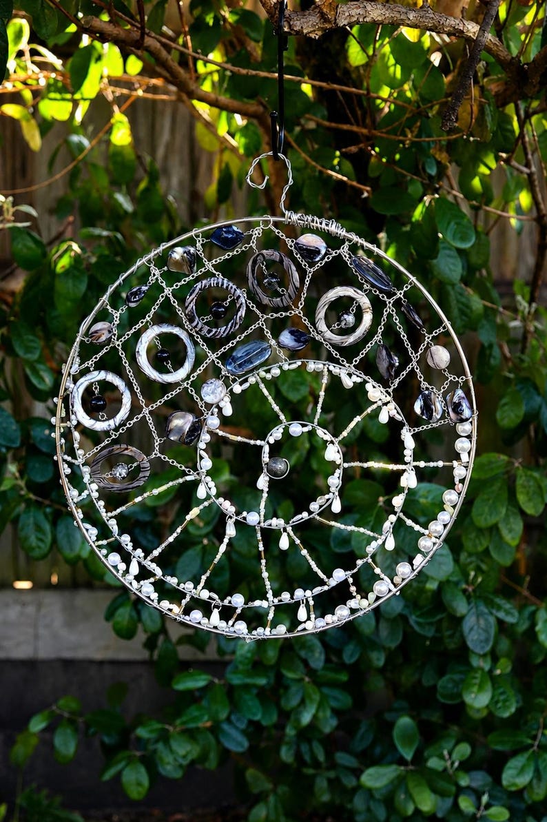 May include: A circular wire wall hanging featuring a spiral design. The structure is adorned with various white, grey, and black beads, along with several circular rings and dark stone-like accents integrated into the silver-toned wire frame.