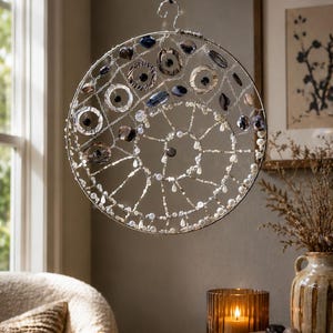 May include: A circular hanging ornament featuring a silver wire frame in a spiral pattern. The structure is adorned with various beads, including dark blue, grey, and clear glass stones, along with small white pearls and metallic accents.