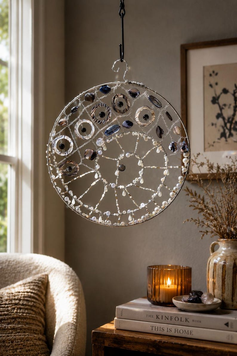 May include: A circular hanging ornament featuring a silver wire frame in a spiral pattern. The structure is adorned with various beads, including dark blue, grey, and clear glass stones, along with small white pearls and metallic accents.