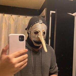 May include: A theatrical mask with a long, curved nose, a white face, and a black hood. The mask has metal accents and a zipper-like mouth. The person is wearing a gray hoodie.