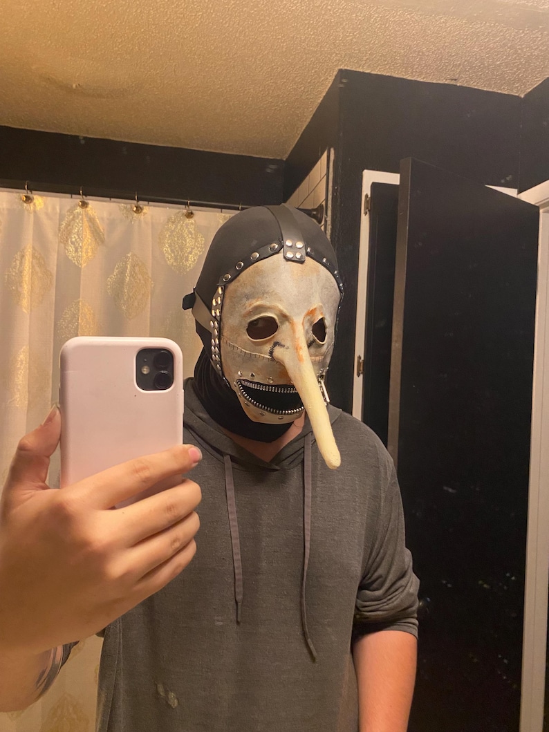 May include: A theatrical mask with a long, curved nose, a white face, and a black hood. The mask has metal accents and a zipper-like mouth. The person is wearing a gray hoodie.