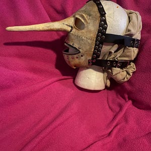 May include: A beige plague doctor mask with a long, curved beak and a zippered mouth. The mask has eye holes and is secured with black leather straps and metal rivets. The mask is displayed on a white foam head against a pink background.