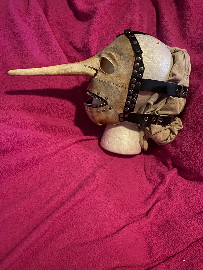 May include: A beige plague doctor mask with a long, curved beak and a zippered mouth. The mask has eye holes and is secured with black leather straps and metal rivets. The mask is displayed on a white foam head against a pink background.