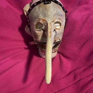 May include: A grotesque mask with a long, pointed nose and stitched details. The mask features a zipper mouth and a leather strap with metal studs. The color palette includes off-white, brown, and black, creating a disturbing aesthetic.