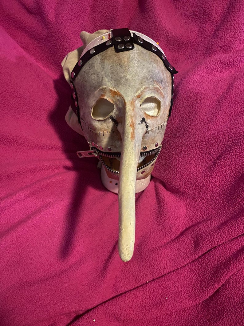 May include: A grotesque mask with a long, pointed nose and stitched details. The mask features a zipper mouth and a leather strap with metal studs. The color palette includes off-white, brown, and black, creating a disturbing aesthetic.