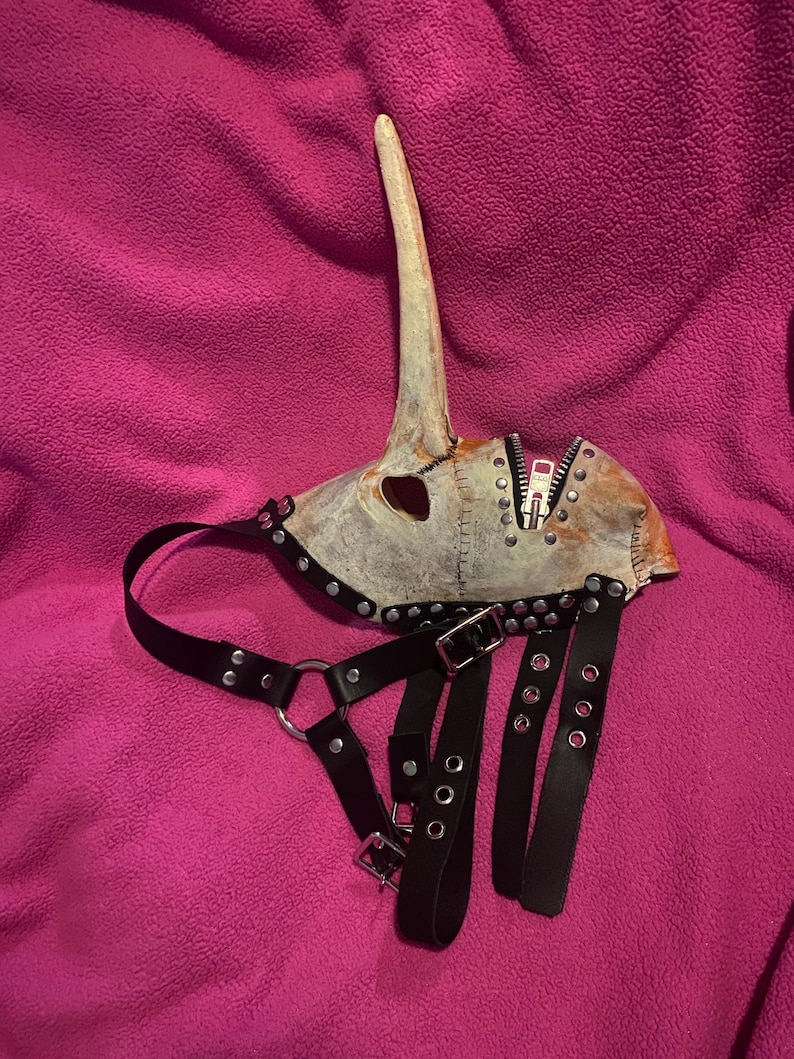 May include: A distressed, off-white mask with a long, curved beak and a zipper detail. The mask is adorned with metal studs and rivets. Black leather straps with buckles and metal rings are attached, suggesting a harness-like design. The mask rests on a pink surface.
