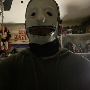 May include: A white mask with a zipper mouth, metal accents, and a black neck covering. The mask has eye holes and a neutral expression. The mask is likely for costume or cosplay purposes.