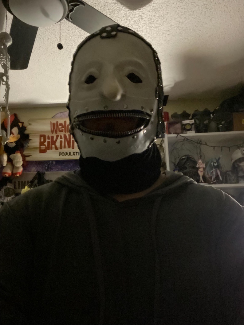 May include: A white mask with a zipper mouth, metal accents, and a black neck covering. The mask has eye holes and a neutral expression. The mask is likely for costume or cosplay purposes.