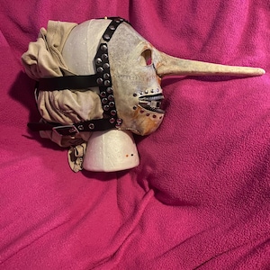 May include: A grotesque mask with a long nose, painted in off-white and tan hues. The mask features a hinged mouth, black leather straps with silver studs, and a fabric head covering. The mask is set on a white foam base against a pink background.