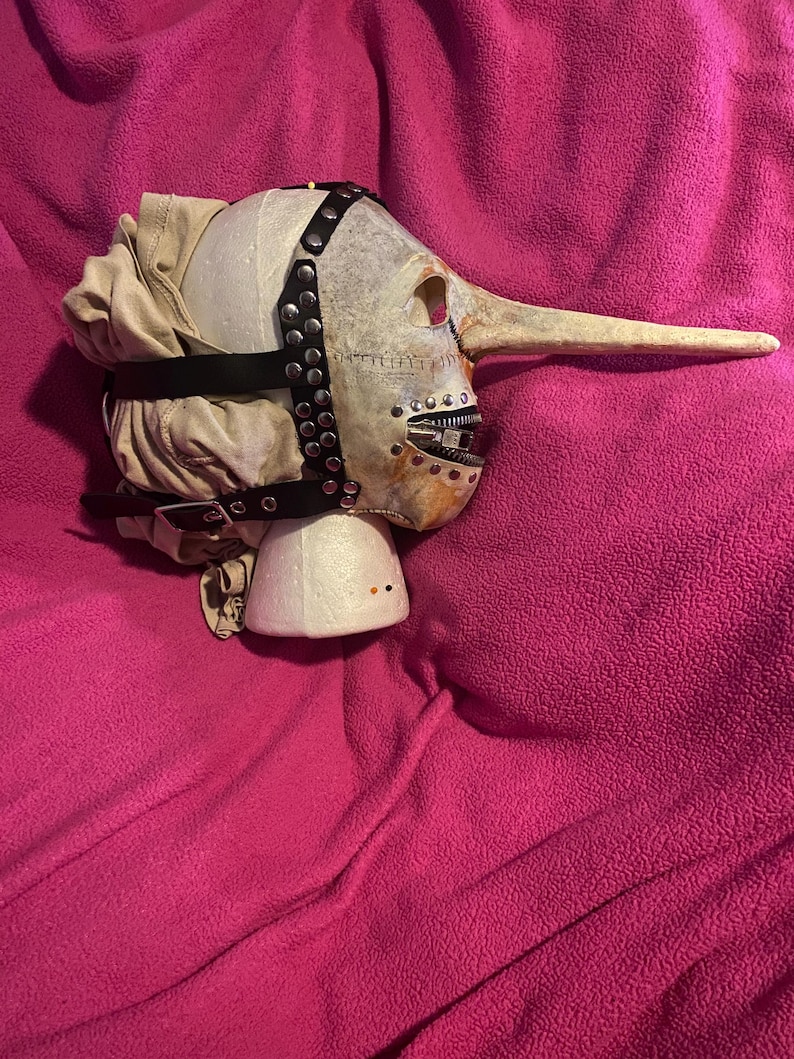 May include: A grotesque mask with a long nose, painted in off-white and tan hues. The mask features a hinged mouth, black leather straps with silver studs, and a fabric head covering. The mask is set on a white foam base against a pink background.