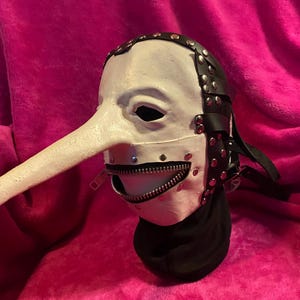 May include: A white mask with a long, curved nose and a zipper mouth. The mask has black leather straps with silver rivets and a black fabric neck covering. The mask is set against a pink background.