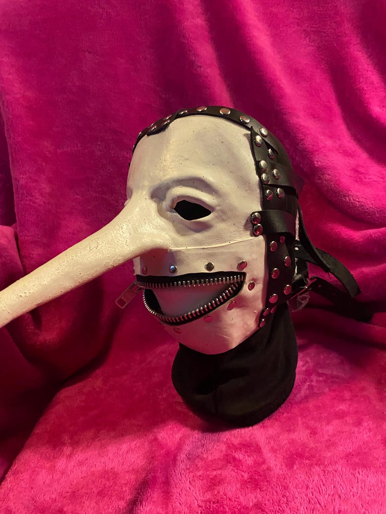 May include: A white mask with a long, curved nose and a zipper mouth. The mask has black leather straps with silver rivets and a black fabric neck covering. The mask is set against a pink background.