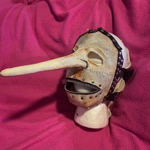 May include: A grotesque mask with a long, pointed nose and a zipper mouth. The mask is painted in a pale, aged color with visible stitching and metal rivets. A black strap with silver studs encircles the head.