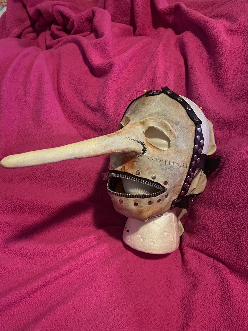 May include: A grotesque mask with a long, pointed nose and a zipper mouth. The mask is painted in a pale, aged color with visible stitching and metal rivets. A black strap with silver studs encircles the head.