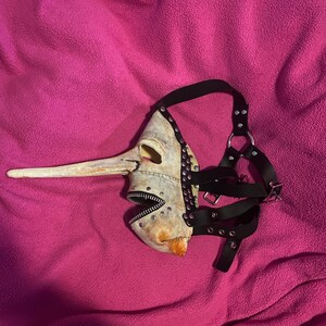May include: A unique, handmade mask with a long, beak-like nose and a zipper mouth. The mask is painted in shades of off-white and tan, with black leather straps and silver-colored studs. The mask is designed to be worn and has an unusual, artistic aesthetic.