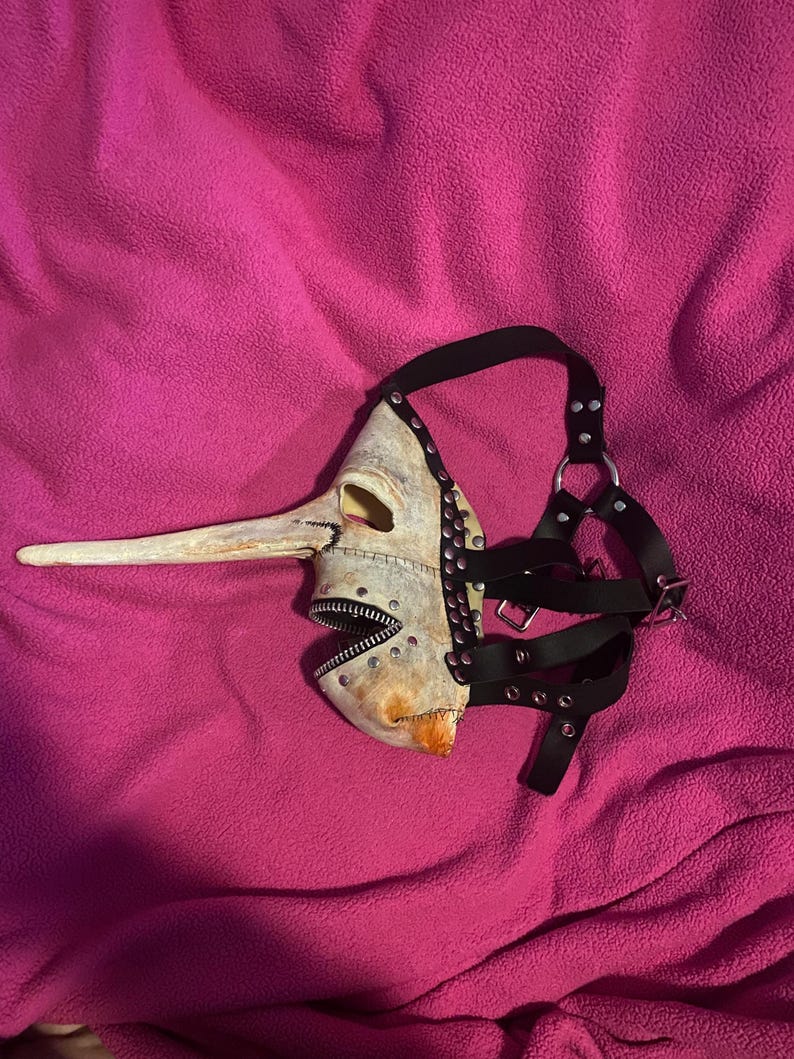 May include: A unique, handmade mask with a long, beak-like nose and a zipper mouth. The mask is painted in shades of off-white and tan, with black leather straps and silver-colored studs. The mask is designed to be worn and has an unusual, artistic aesthetic.