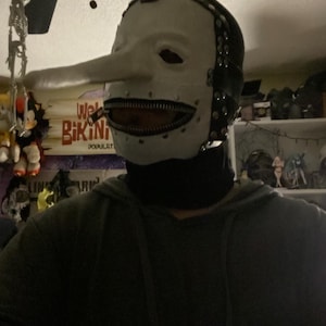 May include: A white mask with a long nose and a zipper mouth. The mask has black accents around the edges and is worn with a black turtleneck. The mask is reminiscent of a character from a horror movie.