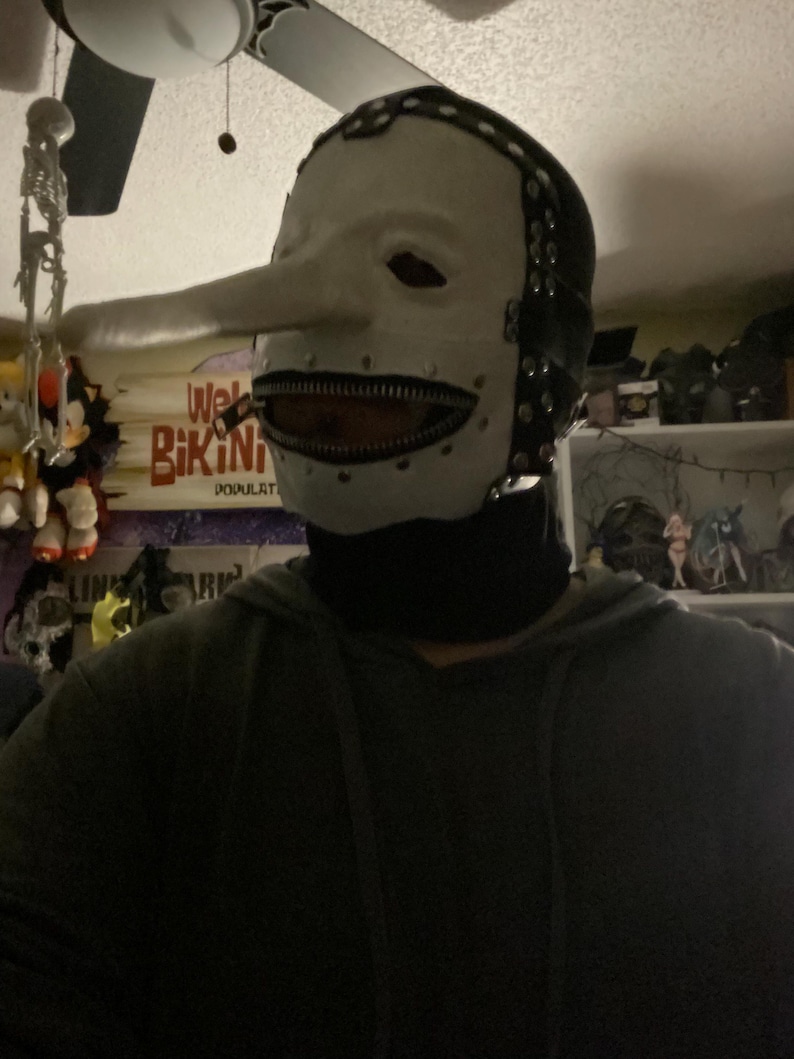 May include: A white mask with a long nose and a zipper mouth. The mask has black accents around the edges and is worn with a black turtleneck. The mask is reminiscent of a character from a horror movie.