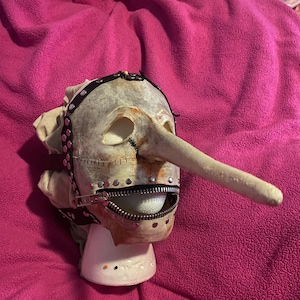 May include: A grotesque mask with a long nose, zipper mouth, and studded leather straps. The mask is off-white with brown and red accents, and is mounted on a white stand. The overall theme is horror or theatrical.