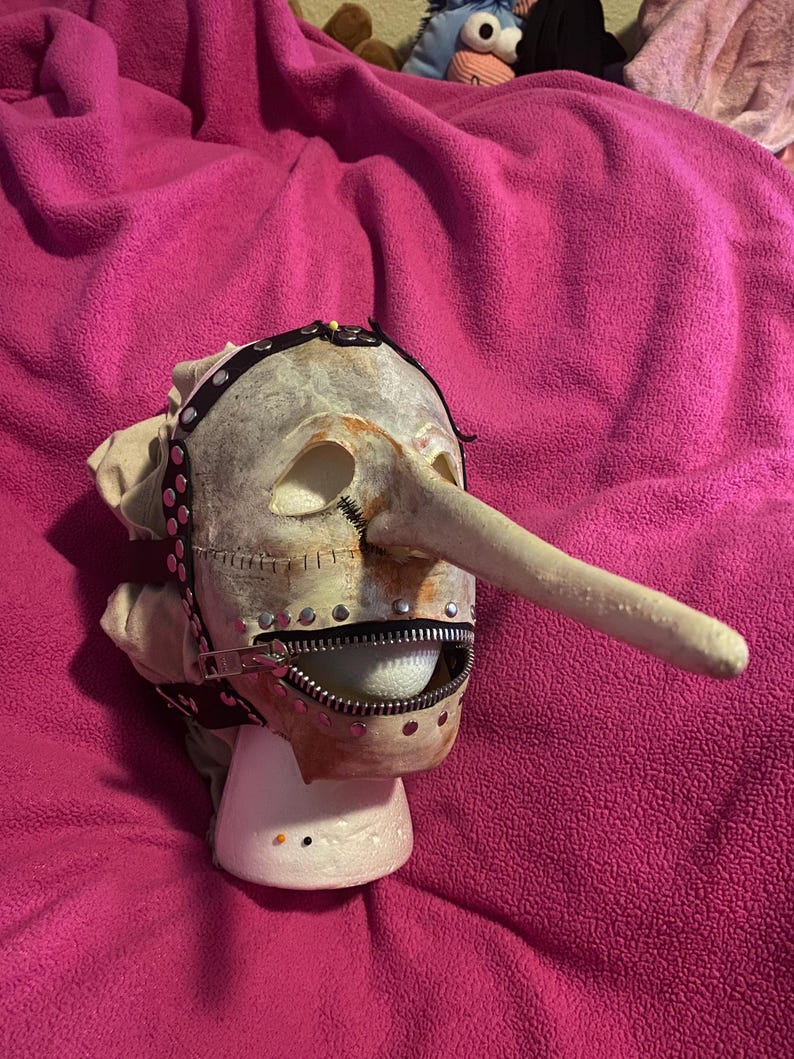 May include: A grotesque mask with a long nose, zipper mouth, and studded leather straps. The mask is off-white with brown and red accents, and is mounted on a white stand. The overall theme is horror or theatrical.