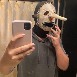 May include: A unique mask with a long nose, zipper mouth, and metal accents. The mask is cream-colored with a black hood and is studded with metal rivets. The mask is being held up in front of a person.