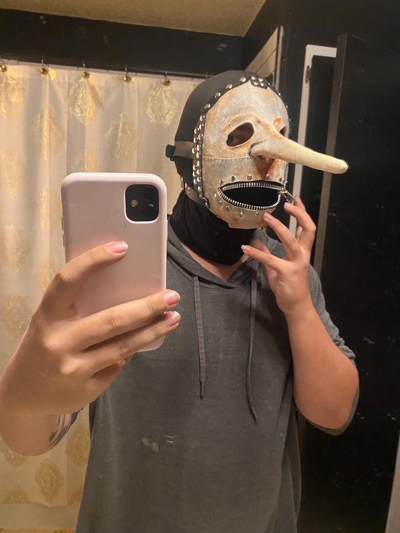 May include: A unique mask with a long nose, zipper mouth, and metal accents. The mask is cream-colored with a black hood and is studded with metal rivets. The mask is being held up in front of a person.
