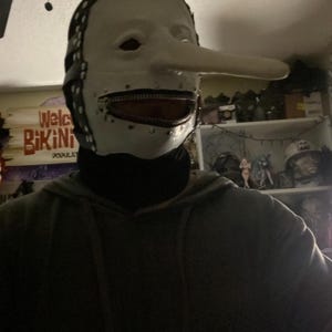 May include: A white mask with a long nose and a zipper mouth. The mask has black leather straps and rivets around the edges. The mask is a costume or prop.