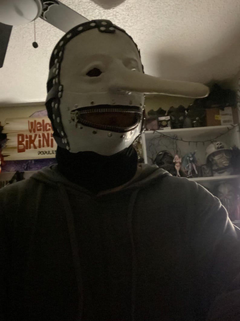 May include: A white mask with a long nose and a zipper mouth. The mask has black leather straps and rivets around the edges. The mask is a costume or prop.