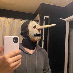 May include: A unique mask with a long nose, reminiscent of a Pinocchio design. The mask features a white face with a zipper mouth, black leather straps, and metal rivets. The mask is worn with a black hood and is part of a costume.