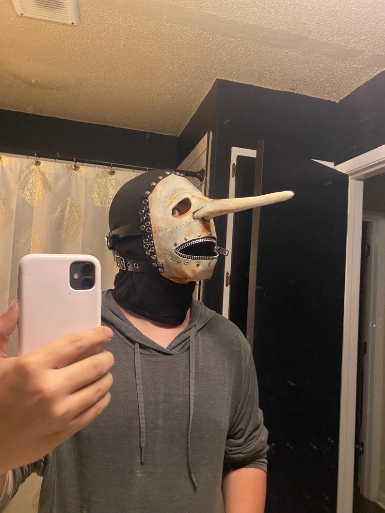 May include: A unique mask with a long nose, reminiscent of a Pinocchio design. The mask features a white face with a zipper mouth, black leather straps, and metal rivets. The mask is worn with a black hood and is part of a costume.