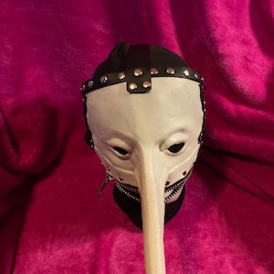May include: A white plague doctor mask with a long, curved beak. The mask has black eye holes, a black leather strap across the forehead, and silver rivets. A zipper runs along the bottom of the beak. The mask is set against a pink background.