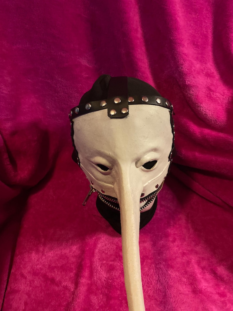 May include: A white plague doctor mask with a long, curved beak. The mask has black eye holes, a black leather strap across the forehead, and silver rivets. A zipper runs along the bottom of the beak. The mask is set against a pink background.