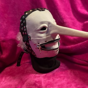 May include: A white mask with a long nose and a zipper mouth. The mask has black leather straps with silver studs and a black neck covering. The mask is set against a pink background.