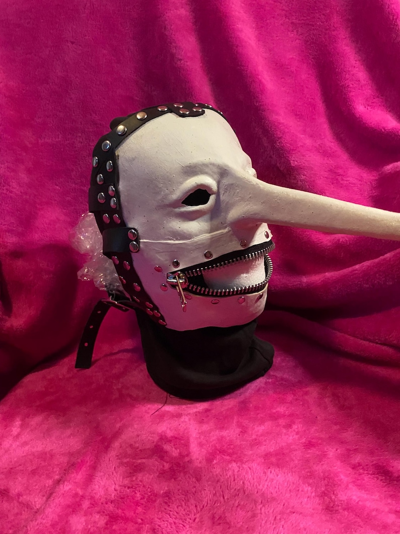 May include: A white mask with a long nose and a zipper mouth. The mask has black leather straps with silver studs and a black neck covering. The mask is set against a pink background.