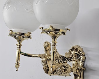 May include: A vintage wall sconce with two frosted glass globe shades. The sconce features a gold-colored metal design with a sculpted figure and ornate detailing. The light fixture is mounted on a white wall.