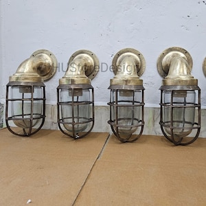 Op de afbeelding: Vier messingkleurige wandlampen met een vintage industriële uitstraling. Elke lamp heeft een ronde basis, een gebogen arm en een glazen behuizing met een kooi. Ze zijn gemonteerd op een witte muur. Geschikt voor binnen en buiten.