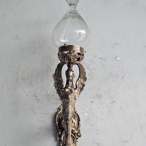 Könnte beinhalten: Eine Wandleuchte mit einem messingfarbenen Metallsockel, einer skulpturalen geflügelten Figur und verziertem Schnörkelwerk. Die Leuchte trägt einen klaren, tropfenförmigen Glasschirm mit facettiertem Abschluss. Der Text https://hustodesign.etsy.com/ ist sichtbar.