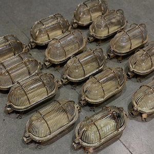 Puede incluir: Una colección de lámparas ovaladas de estilo vintage. Cada lámpara presenta una carcasa de vidrio transparente y acanalado, encerrada en un marco de metal color latón. Las lámparas están dispuestas sobre una superficie gris.