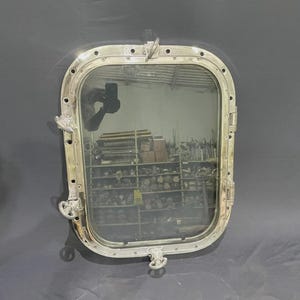 Könnte beinhalten: Ein rechteckiges Bullauge im antiken Stil mit einem silberfarbenen Metallrahmen. Das Fenster hat eine klare Glasscheibe und ist mit Scharnieren und Riegeln befestigt. Das Bullauge ist auf einer dunkelgrauen Oberfläche montiert.