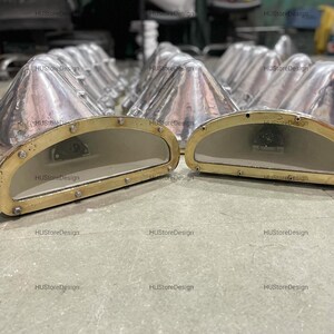 May include: A collection of industrial-style, silver-coloured metal lamps with a brass-coloured trim and a clear glass window. The lamps have a cone-shaped top and a rounded, rectangular window. The lamps are arranged on a grey surface.