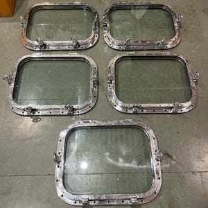 May include: Five rectangular, silver-colored porthole windows with rounded corners. Each window has a clear glass pane and is secured with multiple latches and hinges. The windows are arranged on a gray surface.