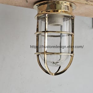 May include: A nautical-style ceiling light fitting featuring a polished brass cage surrounding a clear glass cylindrical shade. The fitting is mounted to a wooden surface and includes the text https://hustoredesign.etsy.com/ printed across the centre.
