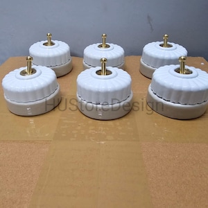 Könnte beinhalten: Sechs weiße Keramik-Lichtschalter mit geripptem Design und Messingwippe. Die Schalter sind auf einer hellbraunen Oberfläche montiert. Die Schalter haben eine Vintage- oder Retro-Ästhetik.