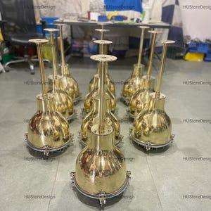 Könnte beinhalten: Eine Sammlung polierter Pendelleuchten aus Messing. Jede Leuchte hat einen abgerundeten Sockel, einen zylindrischen Stiel und eine kreisförmige Montageplatte. Die Leuchten sind in einer Gruppe angeordnet und zeigen ihre reflektierenden Oberflächen und ihr nautisches Design.