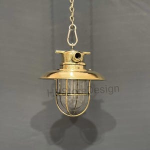 Puede incluir: Una lámpara colgante de color latón con una carcasa de vidrio transparente y una jaula protectora de alambre. La lámpara está suspendida por una cadena y tiene el texto "Hustore Design" en el vidrio.