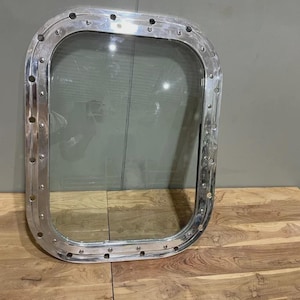 May include: A rectangular, polished silver porthole window with rounded corners. The window has a clear glass pane and is surrounded by a thick, riveted metal frame. The porthole is displayed on a wooden surface.