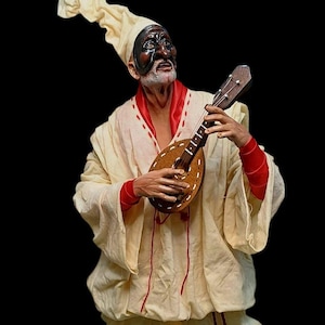 Peut inclure: Figurine d'un musicien vêtu d'une robe blanche avec des bordures rouges, d'un grand chapeau blanc et d'un masque noir. Le musicien joue d'un petit instrument à cordes en bois. Le fond est noir.
