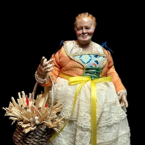 Peut inclure: Figurine détaillée d'une femme en costume traditionnel, tenant un panier tressé rempli de paille et de petits accents rouges. Elle porte une veste orange, une jupe en dentelle blanche et un ruban jaune. La figurine a une expression souriante.